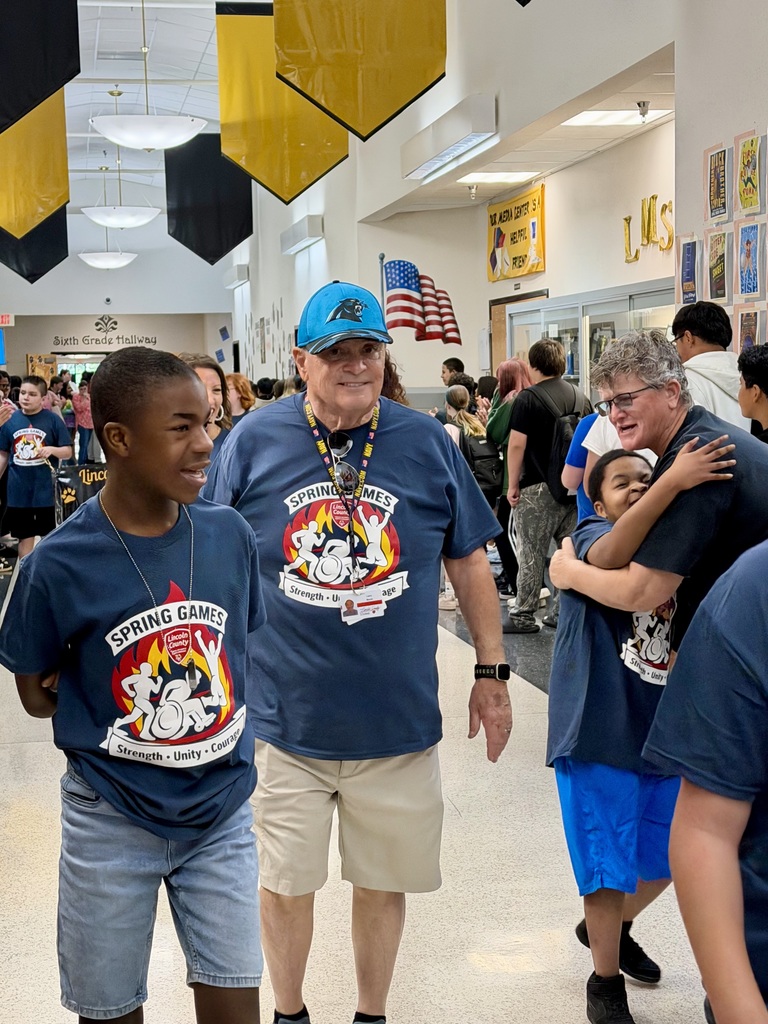 A candid moment in a school hallway where a staff member wearing glasses and a navy blue Spring Games t-shirt shares a hug with a smiling young student. In the foreground, another student walks past, and in the center, a man in a blue Carolina Panthers hat and a navy blue Spring Games t-shirt looks toward the camera. Gold and black banners hang from the ceiling in the background.