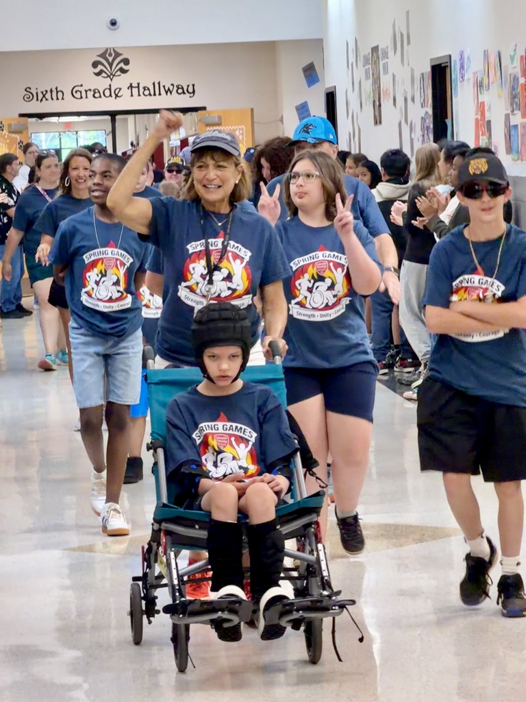 A group of students and staff march down a school hallway under a sign that reads Sixth Grade Hallway. In the center, a staff member in a navy blue Spring Games t-shirt and a grey baseball cap smiles and raises a fist in celebration while pushing a student in a specialized mobility stroller. The student in the stroller is wearing a black protective helmet and a matching navy blue Spring Games t-shirt. Other students walk alongside them, with one student on the right making peace signs for the camera.