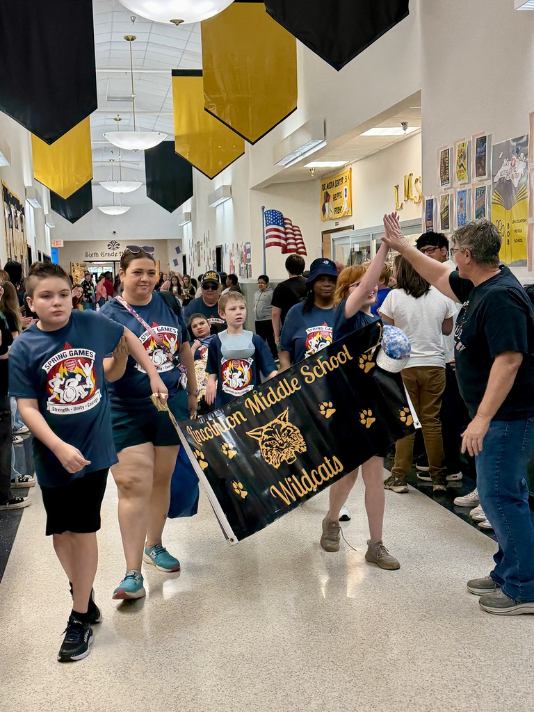 Students and staff from Lincolnton Middle School walk through a school hallway lined with spectators. Two students in the front lead the group by carrying a large black banner with gold lettering that reads Lincolnton Middle School Wildcats, featuring a wildcat logo and paw prints. All participants are wearing navy blue t-shirts with a Spring Games logo that includes the words Strength, Unity, Courage. A staff member on the right is giving a high-five to a student.