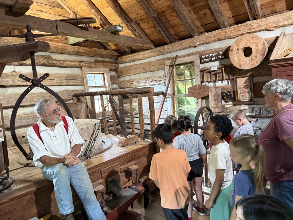 A group of children listens to a man in an old mill. Wooden beams, tools, and equipment are around.