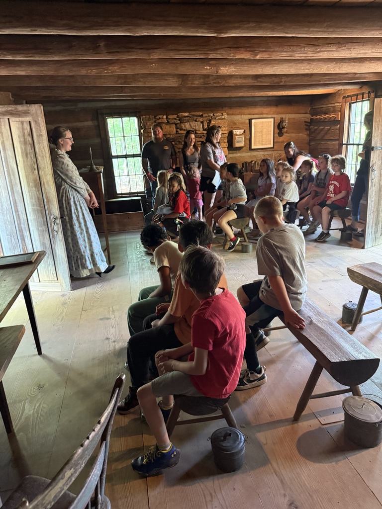 A group of children and adults gather in a log cabin. They sit on benches and a chair.