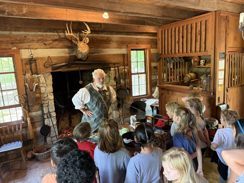A man stands in a log cabin with children, explaining something to them near a fireplace.