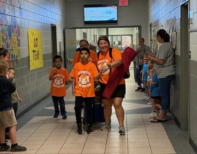 Adults and children wearing orange shirts walk in a hallway with tiled floors and yellow posters.