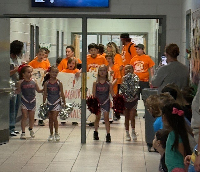 Cheerleaders in matching uniforms walk down a school hallway, holding pom-poms, with a monitor on the wall.