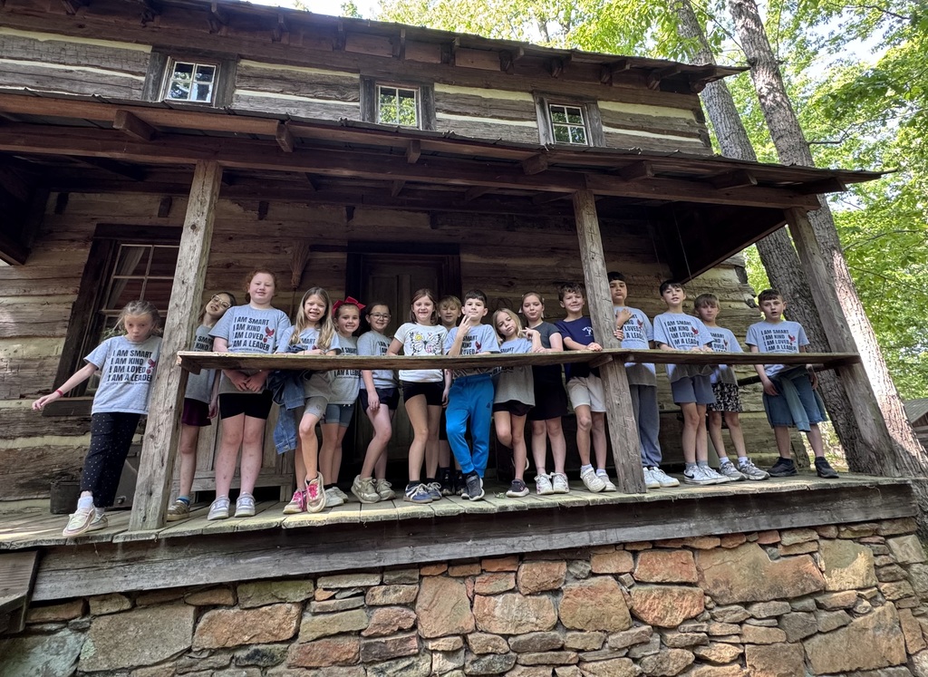 A group of children standing on a porch of a log cabin, with a stone wall at the base.