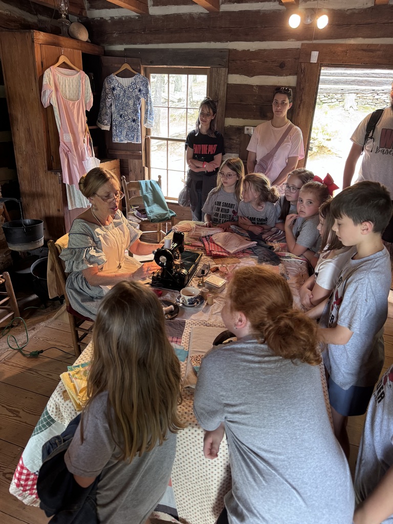 People are gathered around a table with a sewing machine in a room with wooden walls.