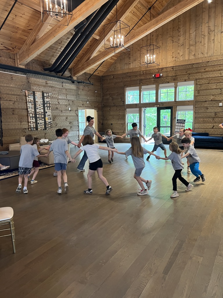 A group of children in a room with wooden floors and wooden beams, dancing in a circle.