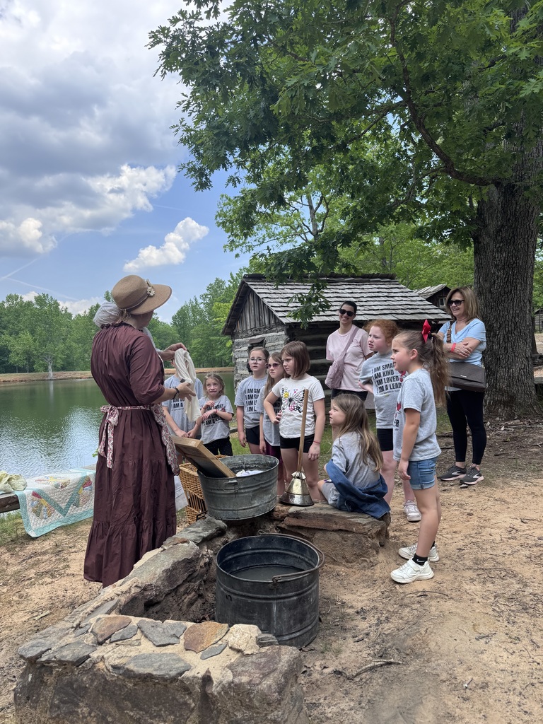 A woman demonstrates hand washing to a group of children in period dress near a rustic fire pit.