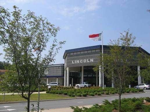 A building named Lincoln has a flag on a pole. Trees and shrubs line the front. Cars are parked in the driveway.