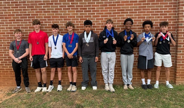 Eleven boys, wearing medals, stand in a row outside a brick wall. They are posing for a photo.