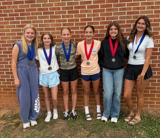 A group of six young girls stands outside a brick wall, all wearing medals around their necks.