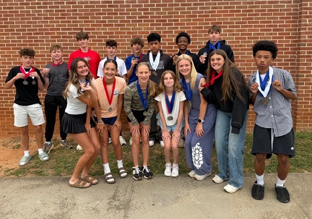 A group of young people wearing medals poses in front of a brick wall. They are smiling.