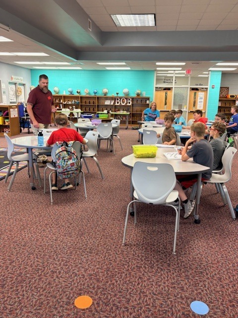 A classroom with tables and chairs, a man standing near shelves with globes, and children seated at tables.