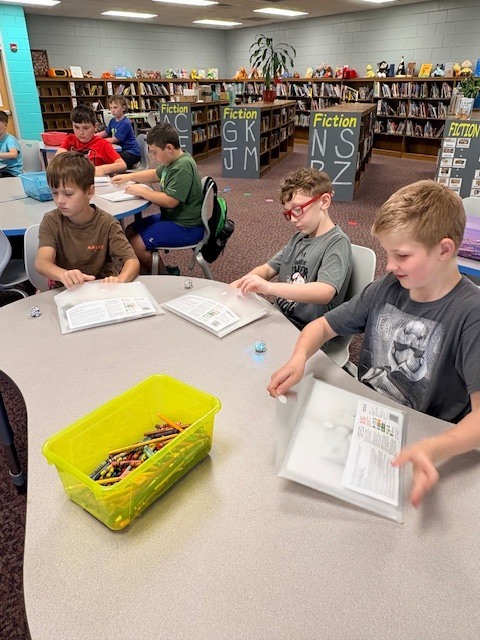 Four children seated around a table, engaged in a reading activity. One has a box of crayons. Background features shelves with books.