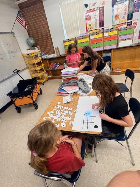 Four girls at a table in a classroom. They have puzzles and paper spread out on the table.