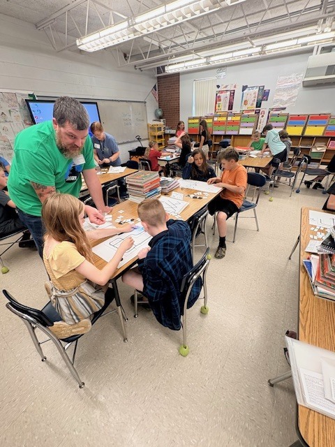 Man in green shirt helps young children seated at desks in a classroom with books and papers.
