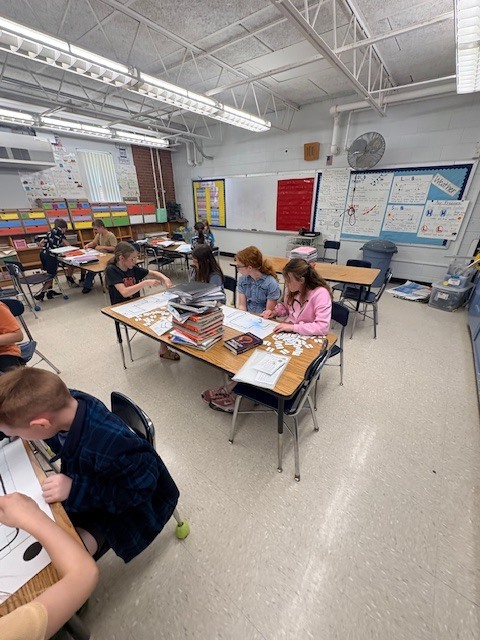 A classroom with students seated at desks, working on projects. Some use books and papers. A white floor and fluorescent lighting are present.