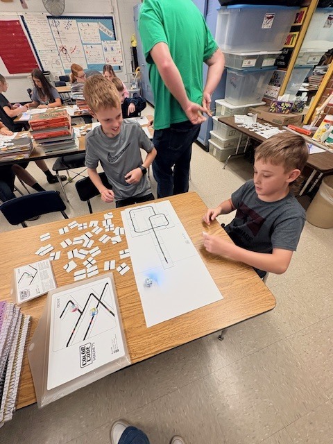 Three boys work at a table with papers and tiles, one points at a paper. Others sit at tables in the background.