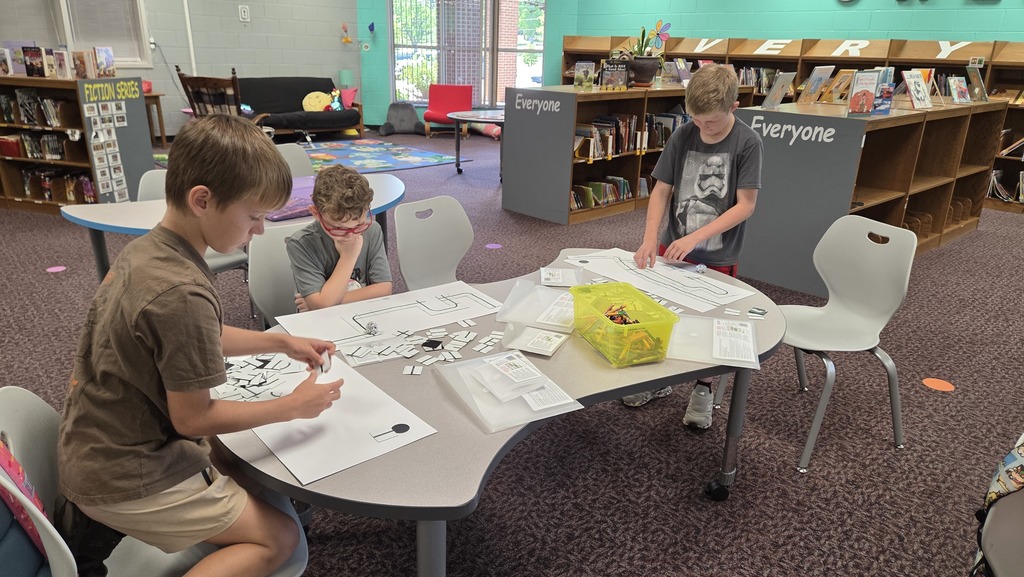 Three boys at a table in a library, engaged in activities with papers and items on the table.