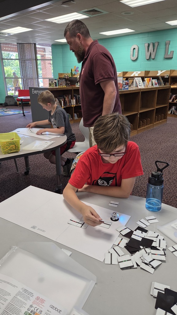 Three people sit at a table in a library, two children focus on a puzzle and a man stands behind them.