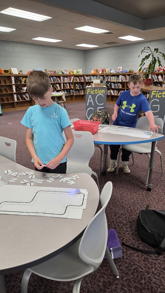 Two children work on a puzzle at a table, surrounded by chairs and bookshelves in a library.