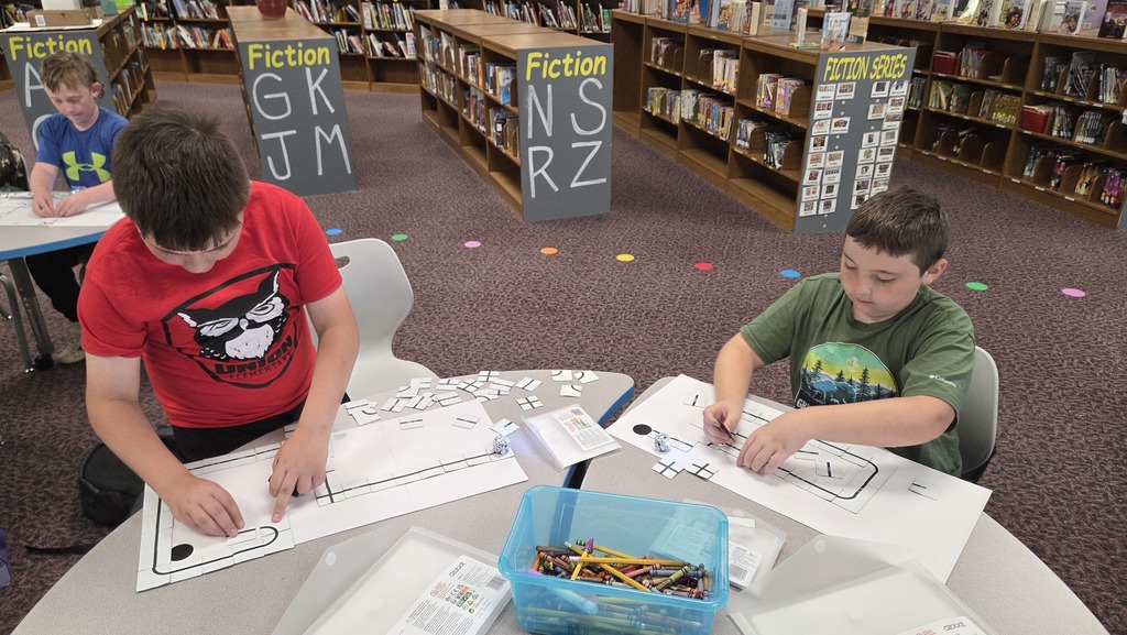 Two boys in a library work on a puzzle on a table, with shelves of books in the background.