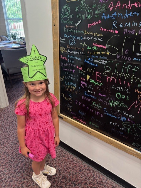 A young girl in a pink dress and green paper hat stands in front of a blackboard with chalk writing.