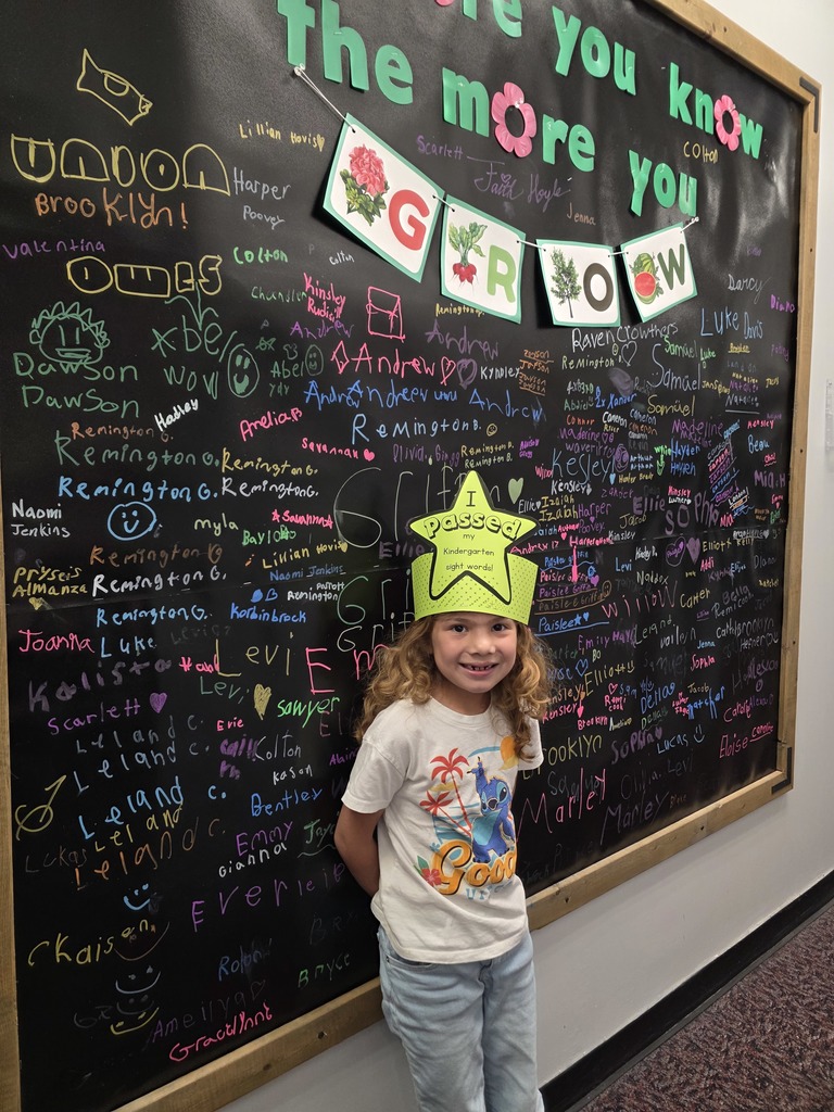 A young girl in a yellow hat poses in front of a chalkboard covered in writing.