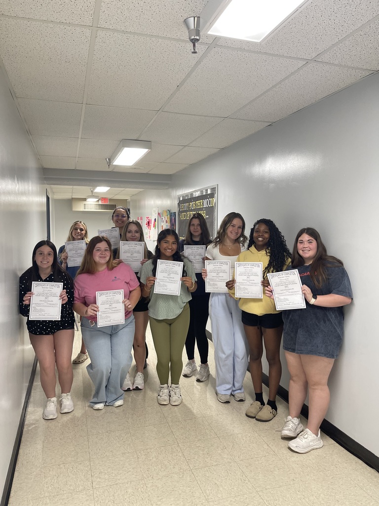 A group of people stand in a hallway, holding white papers, likely certificates. They wear casual clothing and sneakers. The hallway has white walls and a ceiling.