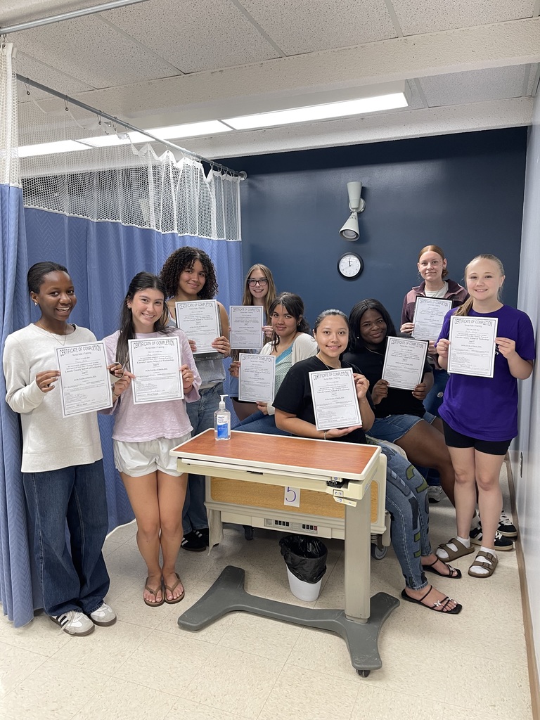 A group of people holding certificates in a hospital room with blue curtains. A table and clock are visible.