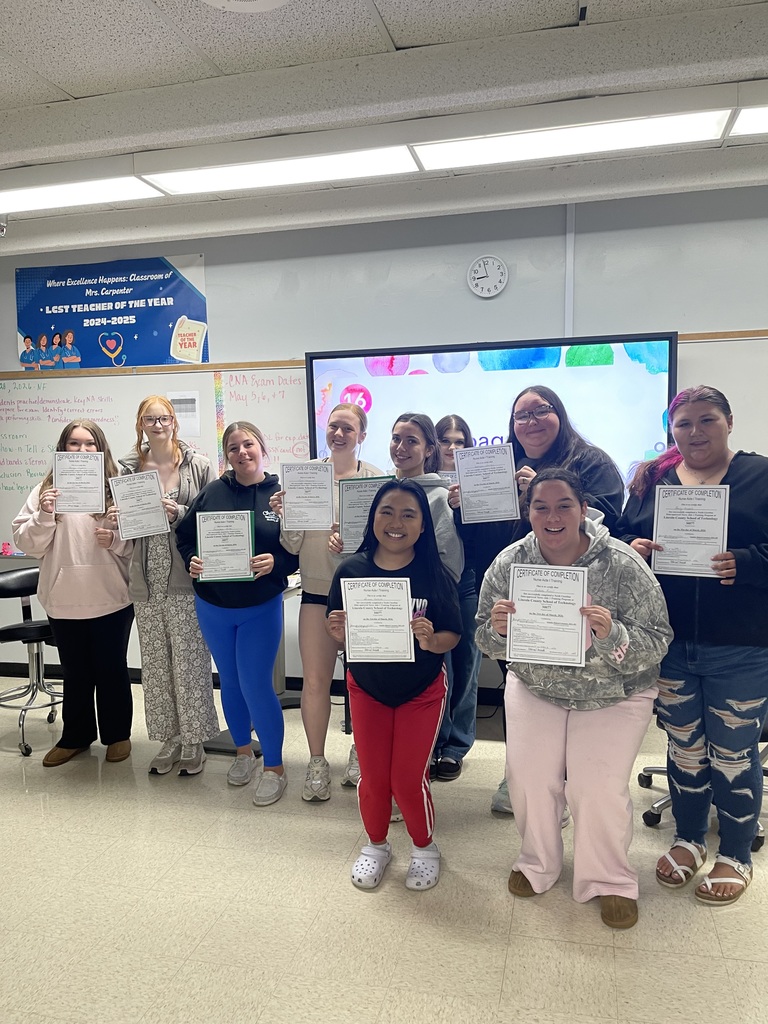 A group of students poses in a classroom, holding certificates, with a screen and whiteboards in the background.