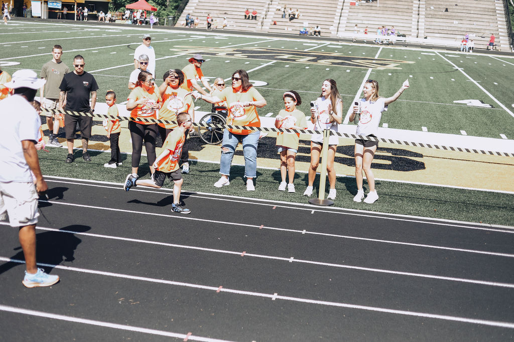 People in orange vests and sneakers stand on a track, watching children run. The track is surrounded by a football field.