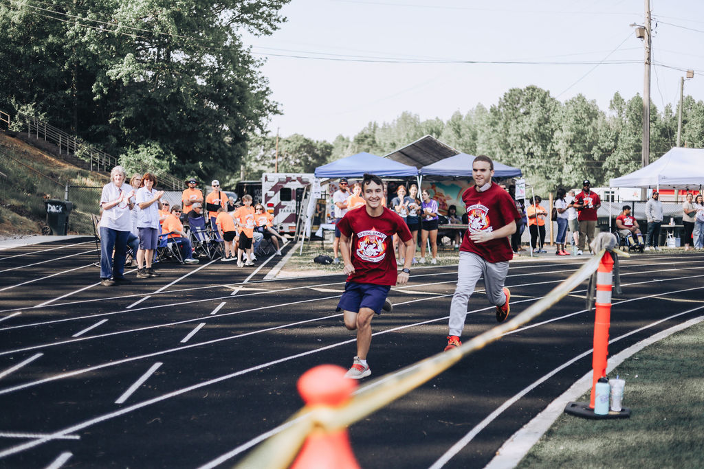 Two runners compete on a track with spectators, tents, and a traffic cone. Trees and a hillside are in the background.
