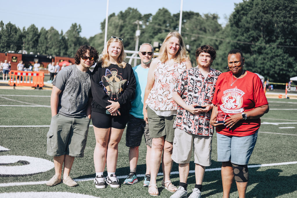 Six people stand on a sports field holding an award; some wear glasses. A white 'S' is on the field. Trees and people are in the distance.