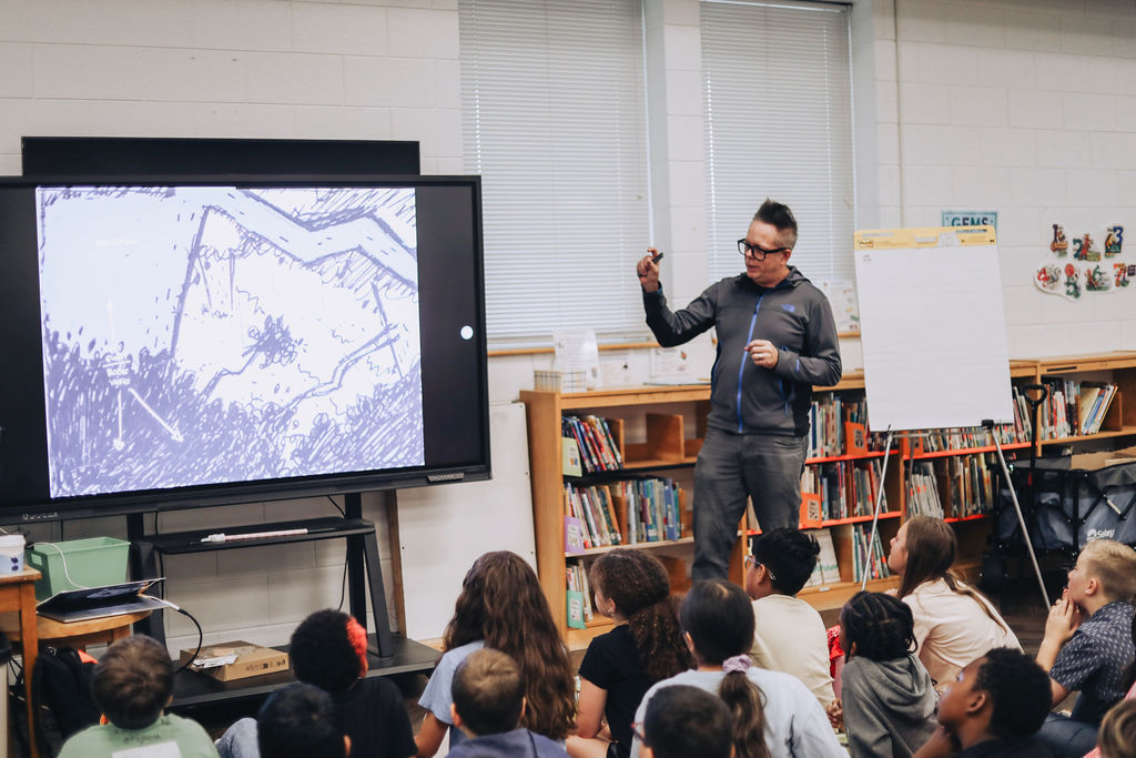 Man speaking to a group of students in a classroom. He stands before a large screen showing a map.