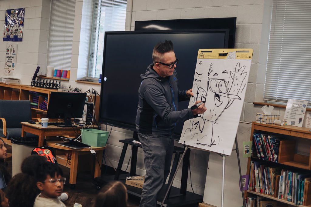 A person draws on a whiteboard in front of a classroom. A television is behind them. Children sit in front.