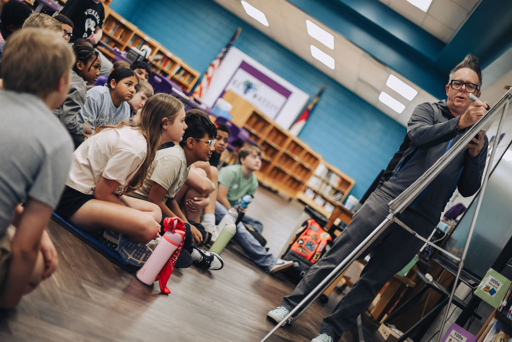A classroom with a man standing before a whiteboard, teaching a group of children sitting on the floor.