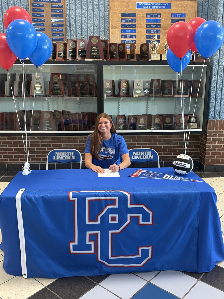 A woman sits at a table with a table cover that reads "FC". She signs a document in front of balloons and trophies in the background.