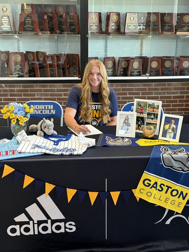 A woman sits at a table with a black backdrop, signing papers. She has a blue shirt and long blonde hair. A table displays Adidas branding and a banner that reads Gaston College.