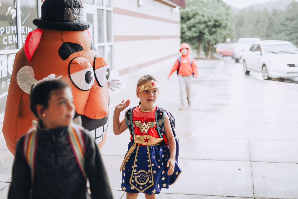 A young girl in a Wonder Woman costume and a boy in a black shirt walk past a pumpkin mascot near a building.