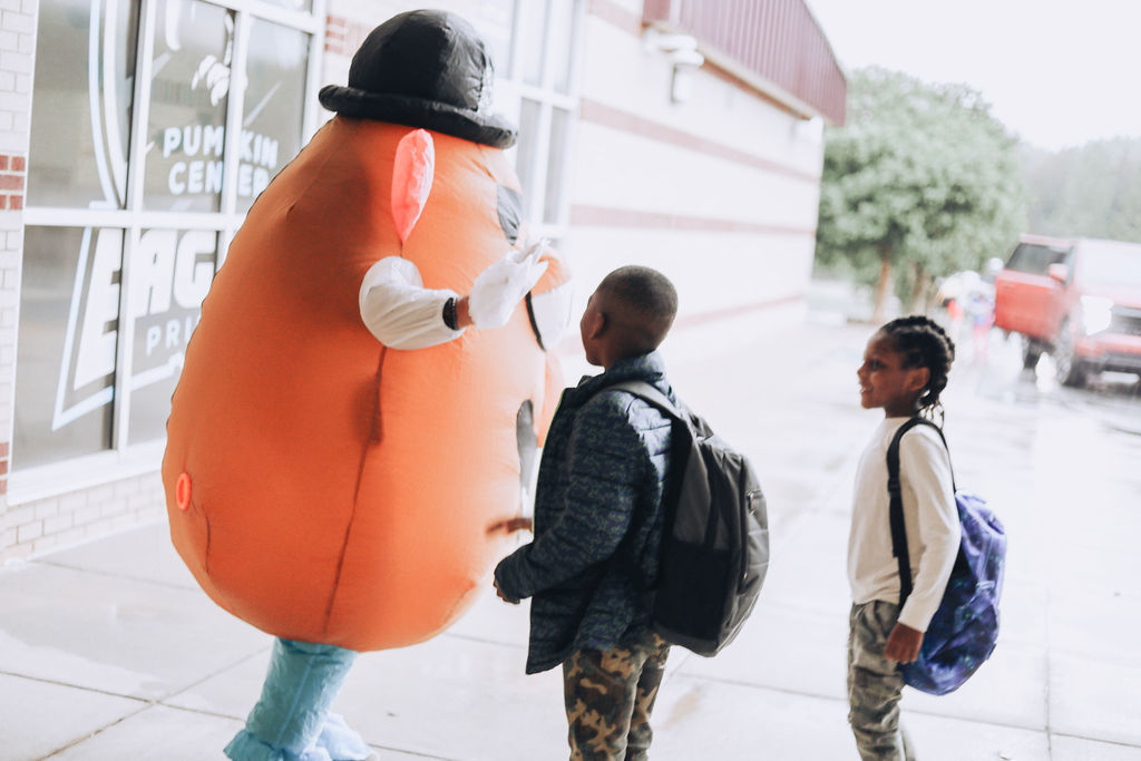 A large mascot resembling a potato with a hat greets two children on a sidewalk. They stand near a building.