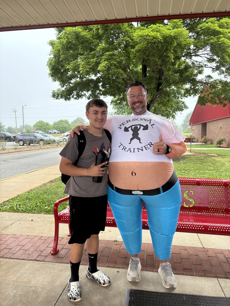A man in a large belly balloon stands next to a boy in front of a red bench.