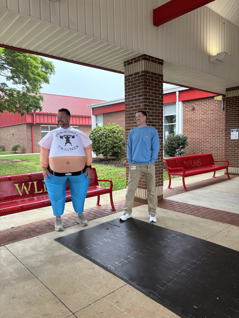 Two people stand in front of a building with a red roof, near red benches. One wears a white shirt, blue pants, and a belt. The other wears blue long-sleeves, tan pants, and sneakers.