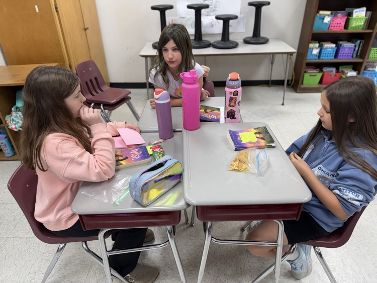 Three young female students sit around a cluster of desks in a classroom setting. The student on the left wears a peach-colored sweatshirt and looks toward the center. The student in the middle, wearing a white t-shirt, sits behind the desks and looks toward the left. The student on the right wears a blue "Stitch" themed hoodie and is looking down at a snack. On the desks are several water bottles, books titled "Wish," a holographic pencil case, and snacks in plastic bags. In the background, there is a white table with black stools placed on top of it and a wooden bookshelf filled with colorful bins..