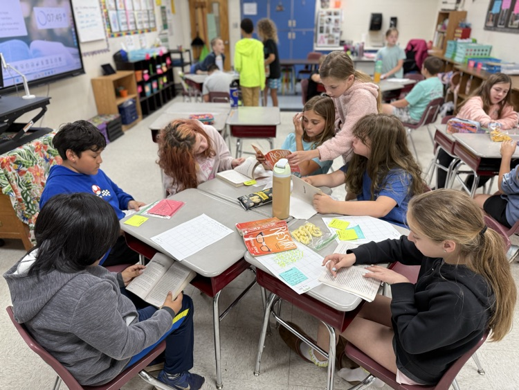 A group of diverse elementary students are gathered around a pod of four desks in a brightly lit classroom, engaged in a collaborative reading activity. Several students are holding and reading copies of the book "Restart" by Gordon Korman. Papers with grids, sticky notes, and snacks are spread across the desk surfaces. In the background, other students are visible working at desks or standing, and a large interactive whiteboard displays a timer and educational content. The atmosphere appears focused and interactive.