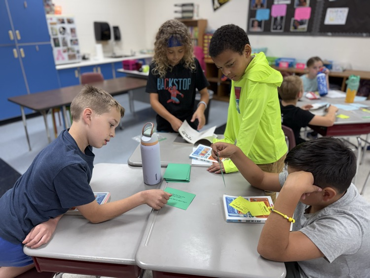 Four male elementary students are engaged in a small group activity at their desks in a classroom. One student on the left points to a green card on the desk while talking to a classmate sitting across from him. A student in a bright neon yellow hoodie and another in a black graphic t-shirt stand behind the desks, looking down at the materials. Books titled "Save Me a Seat" and several green task cards are spread across the workspace along with a purple water bottle. The background shows a classroom environment with blue cabinetry and a bulletin board.