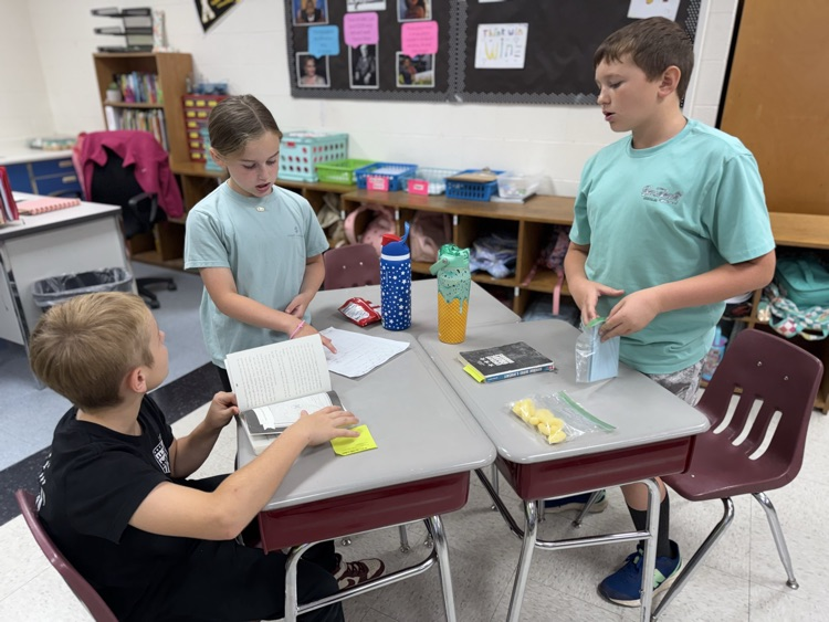 Three elementary students are working together at a group of desks in a classroom. A female student in a light blue t-shirt stands and points to a paper on the desk while speaking to a male student sitting on the left who is holding an open book. On the right, another male student in a light blue t-shirt stands and holds a small blue item. The desks hold various items including water bottles, a book, a small bag of snacks, and a yellow sticky note. In the background, there is a bulletin board with various papers and a wooden shelf with colorful bins. 