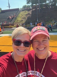 Two smiling women, wearing sunglasses and red shirts, pose for a selfie at a football field.