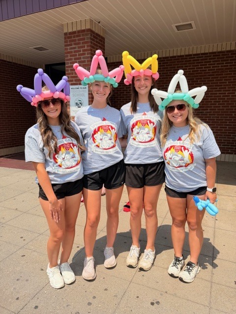 Four women in matching t-shirts and shorts stand in a row, wearing balloon crowns, smiling for the camera.