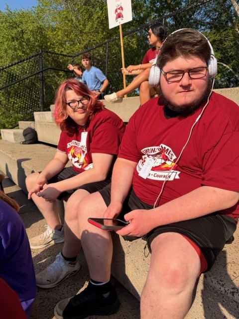 Two people wearing red shirts and black shorts sit on concrete steps. One wears headphones and holds a phone. Behind, another person holds a sign.